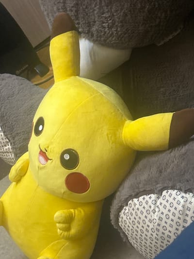 A large yellow Pikachu plush toy is the central subject of the image, resting on a surface covered with soft, textured grey and dark grey materials, likely blankets or cushions. The plush toy is positioned with its head towards the right side of the frame, displaying its signature features: black eyes with white reflections, prominent red circular cheeks, and a small red tongue playfully extended. Its ears are yellow with distinct brown tips, and its limbs are visible.
In the upper right section, a white fabric with a repeating blue geometric pattern, including circles and squares, is partially visible. Towards the lower left, a dark object, possibly an electronic device or a piece of furniture, shows partially legible text that appears to be "KATION". Adjacent to this dark object is a lighter brown or beige surface. The lighting suggests an indoor setting, possibly under artificial light. The image captures a static scene of the plush toy, conveying a playful or comforting mood. No specific activities are taking place, and no direct visual cues to the location of Pernik, Bulgaria, are present in the image itself.