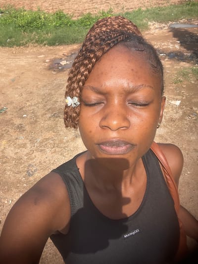 The media file is an image of a young Black woman lying on her back outdoors. She has her eyes closed and appears relaxed. Her hair is braided and adorned with a small, sparkling hair clip shaped like a crown with a flower. She is wearing a black tank top with a thin strap visible over her shoulder, and a brown strap from a bag is also visible. The background is an outdoor scene with a dirt ground, some scattered debris, and patches of green grass. The lighting suggests it is daytime, likely sunny, with shadows visible on the ground and her clothing. There is faint text on her tank top that reads "MeetingGigi". The overall impression is of a serene moment captured outdoors.