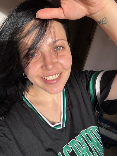 A selfie of a woman is taken indoors. The woman has black hair, fair skin, and green eyes. She is wearing a black shirt with green and white trim. There's a tattoo on her wrist. The photo is rotated 90 degrees counterclockwise, and there is no discernible evidence to indicate a specific location, including Pernik, Bulgaria.
