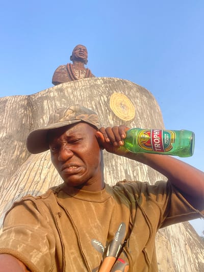 A person in a brown outfit holds a green "Trophy" beer bottle against their forehead in front of a large, grey, elephant-like sculpture with a wood grain pattern. The sky is clear blue. The person wears a camouflage cap and smiles with closed eyes. A small sculpture of a person with a beaded necklace is attached to the side of the larger sculpture. There are also painted objects behind the subject.