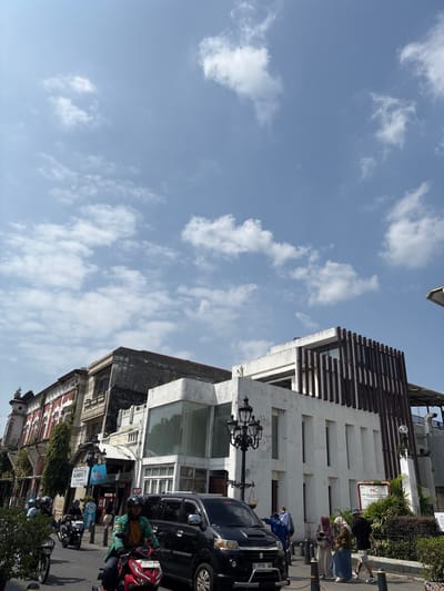 This is a street scene in Semarang, Indonesia, likely in the Kota Lama area given the historical architecture. The image captures a bright, sunny day with a blue sky filled with scattered white clouds. In the foreground, a black car and several motorcycles are on a paved road. One motorcyclist in the foreground wears a green jacket and a black helmet, riding a red motorcycle. Pedestrians, some wearing headscarves and face masks, are visible on the sidewalk.

The buildings lining the street showcase a mix of architectural styles. To the left, an older building features a red and white facade with ornate detailing. To the right, a more modern white building with a slatted wooden exterior stands out. Streetlights with decorative tops are present on the sidewalk. Signs are visible on the buildings, including one that reads "SATE GAUL KAMBING" and another with the logo "Le Minerale." The presence of people, vehicles, and commercial establishments indicates an active urban environment.