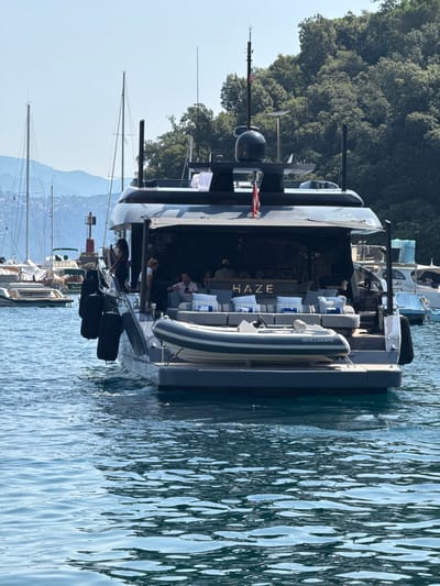 A large, modern motor yacht named "HAZE" is docked in the harbor of Portofino, Italy. The yacht is dark grey with a sleek design. The stern of the yacht features an outdoor seating area with numerous grey cushions and several blue throw pillows. A rigid inflatable boat, also grey and white, is stored on the stern platform, with the name "WILLIAMS" visible on its side. A few people are visible on the yacht, with some seated in the lounge area and others standing near the railing. In the background, other sailboats and smaller boats are moored, and a lush green hillside covered in trees rises behind the harbor. The water in the foreground is a clear blue with gentle ripples. The sky is bright and clear.
