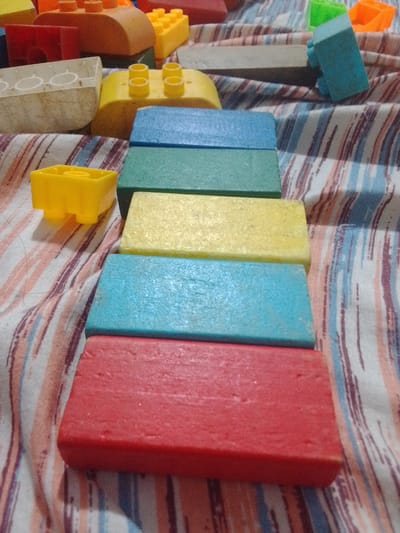 The image displays a collection of colorful building blocks, predominantly red, blue, yellow, and green, arranged on a striped fabric surface. The blocks are of various shapes and sizes, resembling common toy building bricks. One yellow block, shaped like a small house with four studs, is positioned to the left of the frame. To its right, several rectangular blocks are stacked. The bottom-most is red, followed by a blue block, then a yellow block, and finally a green block. In the background, more blocks are visible, including a white one with circular indentations and an orange one. The lighting suggests an indoor setting, possibly a bedroom or play area. There are no people or animals present in the image. The scene is static and shows no activity. The context of La Tagua, Venezuela is not discernible from the visual information in the image.