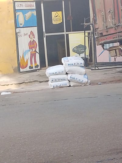 The image depicts a street scene in Maiburiji, Nigeria, with a building entrance and several stacked bags of cement on the side of the road. The building facade features murals: one of "CCTV - CAMERA" with images of security cameras, and another of a "Fire Man" with a fire hose. A yellow "CCTV WARNING" sign is also visible on the door. To the right of the entrance, a bright yellow sign with the text "24/7 Colorful Imaging" and a "ColorVu" logo is partially visible. A large, red shipping container is situated behind the building. A stack of several white bags, labeled "BUA P.O.P", is positioned on the cracked concrete pavement in front of the building. The street in the foreground appears to be asphalt.