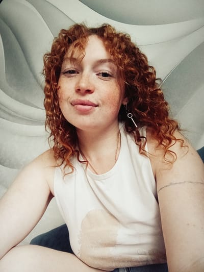 This is a close-up, high-angle portrait of a young adult woman lying down and looking directly at the camera. She has fair skin, a scattering of prominent freckles across her nose and cheeks, and voluminous, curly, reddish-brown hair. Her expression is soft and calm, with a slight, gentle smile and her lips slightly parted.
She is wearing a simple white tank top or t-shirt, and a thin gold-colored necklace is visible around her neck. A distinctive silver-toned earring, composed of a small circular hoop connected to a long, slender bar, hangs from her right earlobe (viewer's left). A dark blue fabric, possibly denim, can be seen near her left side, suggesting she is wearing jeans or similar attire.
The background is an indistinct indoor setting, featuring a light gray or off-white surface with gentle, curved contours, possibly a patterned headboard, sofa, or upholstered furniture. The lighting is soft and even, suggesting ambient light rather than direct sunlight. No specific time of day or weather conditions are discernible from the image. The overall mood is relaxed and intimate. No text is visible. The provided location context for this media file is Yurgamysh, Russia.