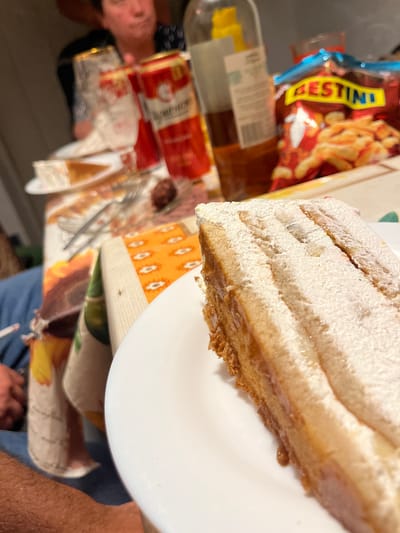 The image depicts a close-up view of a slice of layered cake with powdered sugar dusting on a white plate. In the background, out of focus, are people, a bottle of alcohol, a can of beer, and a bag of snacks. The setting appears to be a casual gathering or meal, likely indoors, given the lighting and arrangement of items on a table. The tablecloth has a colorful pattern. The image is taken at a low angle, focusing on the dessert. There is no information to indicate the location in the city of Dragoevo, Bulgaria.