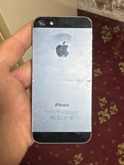 An iPhone 5s is held in a left hand, with the back of the phone facing the camera. The phone is silver and has the Apple logo and "iPhone" printed on the back. The camera and flash are visible at the top of the phone. The phone is positioned over a red carpet with a floral pattern. The background is blurred and appears to be a wall. The image was taken in Kokand City, Uzbekistan.