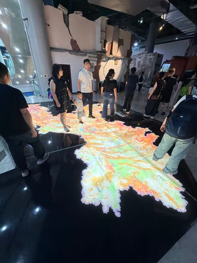 The image depicts an indoor scene with several people observing a large, illuminated map projected onto the floor. The map, which appears to be of a geographical region with varying colorations indicating different features, is centrally located. Several individuals are standing around the map, some looking down at it, while others are in transit.
In the foreground, a person wearing a black t-shirt and dark pants is walking across the floor, partially obscuring the map. To their right, a woman in a black patterned dress and a child in a light-colored shirt and shorts are standing, holding hands and looking at the map. Behind them, a man in a white polo shirt and dark pants is also observing the map. Further back, other individuals are visible, some with backpacks, indicating they are likely visitors in a museum or exhibition space.
The environment appears to be an exhibition hall, with exposed brickwork and structural elements visible on the walls, suggesting a theme related to reconstruction or historical events. The lighting is subdued, with the projected map being the primary light source in its immediate vicinity. The floor is dark and reflective, with some circular light fixtures embedded in it. The overall impression is of an educational or commemorative exhibit.
While the presence of the map suggests a location with geographical significance, the prompt states the location is 江北城街道, China. This information would imply the map is of a region within or related to 江北城街道, though this cannot be definitively determined solely from the visual information in the image.