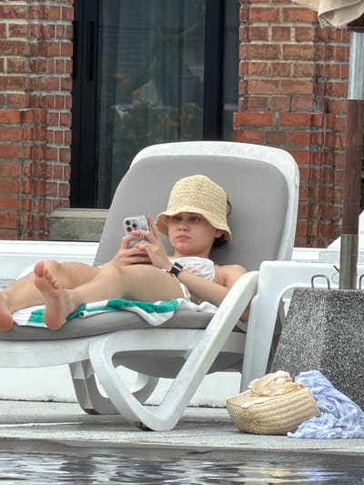 An adult female is sitting on a white lounge chair by a pool, holding a smartphone. She is wearing a straw hat and a white bikini top. A blue and white towel is draped on the chair. Next to the chair, on the ground, is a straw handbag and a blue and white patterned cloth. The background shows a brick wall and a window.