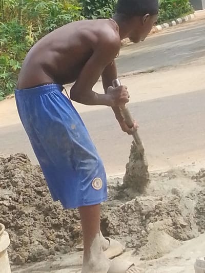 In the city of Alor, Nigeria, a young male, shirtless and with dark skin, is depicted engaged in manual labor. He wears blue athletic shorts, stained with dirt, which feature a circular yellow and black emblem on the lower left leg. Light-colored, dirt-covered sandals are visible on his feet. A light-colored necklace adorns his neck. He is leaning forward, holding a shovel or spade with both hands, actively digging into or lifting a pile of damp, brownish-grey granular material, likely a construction mixture, situated on the ground. The tool's blade is embedded in the material, which forms a significant mound directly in front of him. To the left, the rim of a light-colored bucket is partially visible. The background reveals a paved road with a white and black striped curb on the right, and green foliage extends across the upper left, indicating an outdoor, daylight scene.
