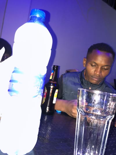 The image shows a young African man sitting at a table in a dimly lit establishment, possibly a bar or restaurant, in Arusha, Tanzania. He is wearing a dark blue vest and a white shirt. He is looking down, possibly at a phone or a drink.  A clear glass sits on the table in front of him.  A bottle of water and a bottle of beer are sitting on the table in front of him. The table is a dark, shiny surface with a pattern. The walls are white, and the lighting is blue.  It is possible that he is waiting for someone, or talking on his phone. The image is taken from a low angle, with the bottle of water in the foreground.  The man's face is visible, and he appears to be in his early 20s.