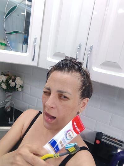 A woman is holding a toothbrush and toothpaste tube in front of her while winking. She is standing in what appears to be a bathroom or kitchen with white cabinets, a sink, and a white brick-patterned backsplash. The toothpaste is a Colgate Total brand. A toaster oven is visible in the background. There is no information to confirm the location of Andreyevo, Russia.