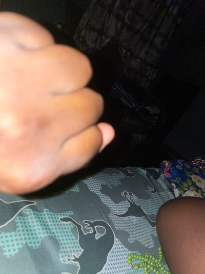 A close-up image shows a person's hand in the foreground. The hand appears to be clenched. In the midground, there is a pattern of stylized dog figures printed on a fabric, with green beads visible on the side. The background is dark, with some undefined objects. The location is Kurunduma, Nigeria.