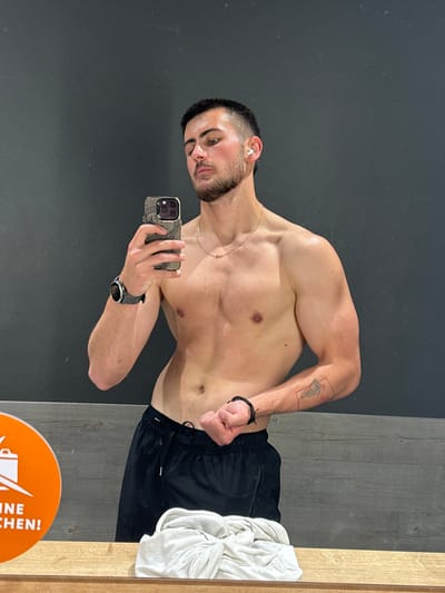 A shirtless man with dark hair and a trimmed beard is posing for a selfie in what appears to be a gym or fitness center. He is holding a smartphone with a decorative case in his left hand, and flexing his right arm to display his bicep. He is wearing black athletic shorts, a wristwatch on his left wrist, a small gold chain around his neck, and a bracelet on his right wrist. A tattoo is visible on his upper right arm.
The background consists of a gray wall on the left and a wood-paneled wall with a mirror on the right. A white towel is draped over the mirror frame. On the floor, there is a circular orange sign with an advertisement or branding on it. The location is a gym, potentially in Essen, Germany, based on the language used on the sign.