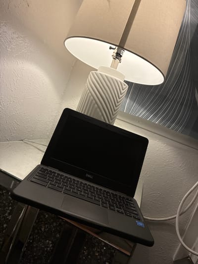 The image shows a Dell Chromebook laptop placed on a reflective surface, possibly a glass-top table. The laptop is open, with its screen facing upwards and appearing to be off or displaying a dark image. The keyboard and touchpad are visible, featuring standard QWERTY layout. A lamp with a neutral-colored shade and a textured base is positioned behind and to the right of the laptop, casting light. The background includes a textured wall and a patterned wallpaper with vertical lines. A white cable is visible running along the wall. The setting appears to be indoors, in a room possibly located in Balch Springs, United States of America. There are no people or animals present in the image.