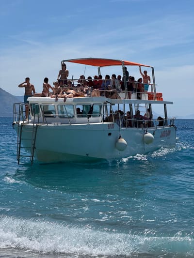 A white boat named "JADRAN" is sailing in the turquoise waters off the coast of Prijevor, Montenegro. The boat is crowded with approximately 20 people, most of whom are young adults, wearing swimwear and enjoying the sunny weather. Some are standing on the upper deck, while others are seated or lying down. The boat has an orange canopy providing shade on the upper deck. The water is clear and choppy, with small waves breaking near the shore. The background shows a mountainous landscape characteristic of the Adriatic coast. The sky is clear and blue.