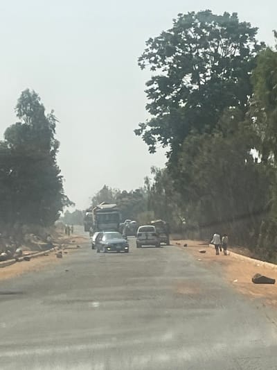 The scene depicts a two-lane paved road in Hoss, Nigeria, under hazy atmospheric conditions. The road recedes into the distance, flanked by dense green foliage and trees. The unpaved shoulders are reddish-brown dirt, littered with scattered rocks and debris. Multiple vehicles are observed moving away from the viewer. In the left lane, a dark sedan with illuminated headlights leads a lighter-colored SUV. Further down the road, a large articulated lorry is visible in the left lane. On the left shoulder, several indistinct figures are present. On the right shoulder, two individuals, wearing light-colored clothing, walk away from the camera. The sky is a uniform light gray.