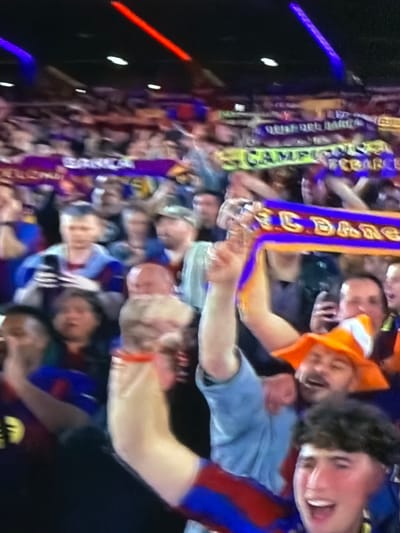 The image shows a crowd of people, likely at a sporting event given the scarves and team colors. Individuals are visible, some smiling and cheering. A man is wearing an orange hood. Scarves with words such as "Campions" and "FC BARCA" are visible. The exact location in Sant Martí Sarroca, Spain is not specified.