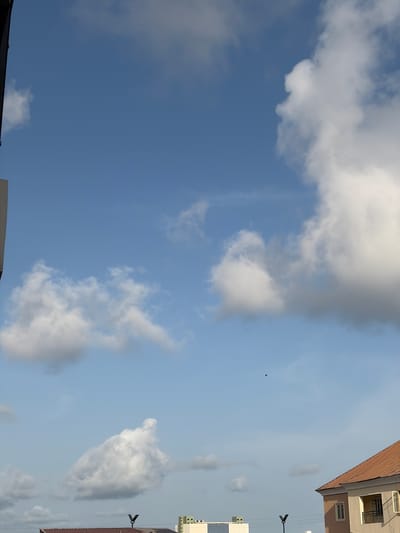 The image shows a daytime sky in Igboefon, Nigeria. A mix of fluffy white cumulus and thin cirrus clouds are scattered across a blue sky. A dark, indistinct bird is visible in the distance. Rooftops and building facades of multistory structures are visible along the right and bottom edges of the frame, indicating an urban environment. Streetlights are attached to buildings.
