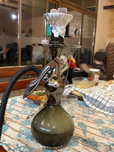 The image depicts a close-up view of a hookah on a table, set within what appears to be a domestic indoor environment. The hookah, made of dark, reflective metal and green glass, is the central subject. It is adorned with aluminum foil at the top, containing what looks like charcoal. A black hose extends from the side, and a pair of metal tongs is attached to the hookah's stem.
On the table, beneath the hookah, a light-colored tablecloth with a nautical-themed pattern of shells and ship wheels is visible. Several objects are arranged on the tablecloth, including a small bowl filled with a reddish food item, a glass, and a jar of preserves. A checkered cloth partially covers a plate with slices of bread. In the background, through glass doors, a dimly lit room is visible, with furniture and various objects that suggest it is a living space. The overall lighting is dim, contributing to a relaxed or evening atmosphere. There are no people visible in the frame. The location is identified as Kamenar, Bulgaria.