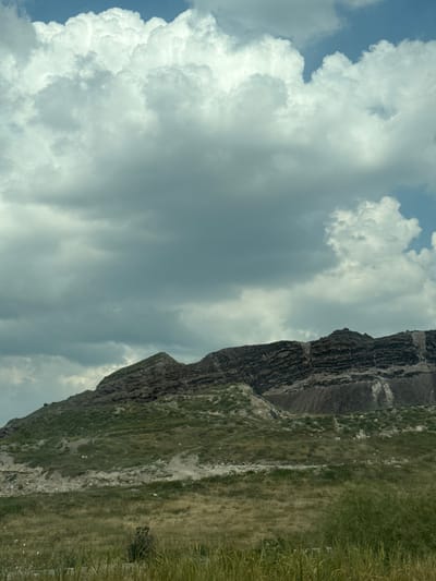 The image depicts a landscape featuring a mountainous terrain with visible layers of rock formations. The foreground is dominated by dry, tall grass. The middle ground shows a green, sloped hillside leading up to the rocky mountains. Scattered white stones are visible on the hillside. The upper portion of the image is filled with a dramatic sky characterized by large, dense, white and gray clouds against a bright blue sky. The scene appears to be located in the vicinity of Arzni, Armenia, suggesting a natural, possibly arid or semi-arid environment. No people or active events are depicted.