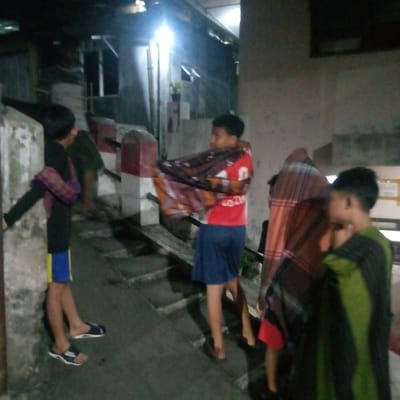 The image depicts an outdoor, low-light scene featuring four young males in a residential alleyway or pathway in the city of Bogor, Indonesia. The environment includes a light-colored concrete wall on the far left, a sloped pathway or steps leading upwards from left to right, and two red-and-white striped bollards positioned centrally. A bright artificial light source illuminates the background, revealing parts of a building structure.
From left to right:
1. A male, with his back partially turned, stands near the concrete wall. He wears a dark long-sleeved shirt, blue and yellow shorts, and black-and-white sandals. A purple and yellow patterned sarong is draped over his left shoulder.
2. A male stands slightly uphill on the pathway, wearing a red t-shirt with a white graphic and blue shorts. He holds a large, multi-colored patterned sarong draped over his shoulders.
3. A male, partially obscured and facing away from the camera, is visible further up the pathway, wrapped in a reddish-brown plaid patterned sarong.
4. A male, partially obscured and facing towards the right edge of the frame, is seen on the far right, wrapped in a green and black striped or patterned sarong.
All individuals are draped in sarongs, indicating a casual, informal evening setting. No specific actions or interactions are discernible beyond their presence in the scene.