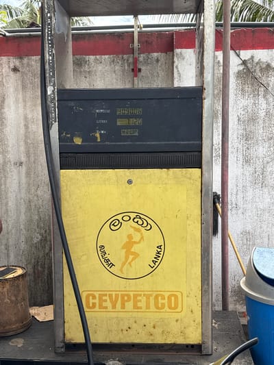 This media file captures a close-up, angled view of a fuel dispenser, likely at a gas station in Ahangama, Sri Lanka. The dispenser is predominantly black and yellow, with a worn, textured surface. The black section features digital displays showing "30900.08" under "RUPEES" and "1.120" under "LITRES," and "434.00" under "Rs/Litre." The yellow section displays a circular logo with text in what appears to be Sinhala and English. Inside the circle is a silhouette of a person, possibly reclining or stretching. The text visible on the yellow panel includes "CEYPEEICO" vertically along the right side, and "LANKA" and a Sinhala script within the circular logo.
The dispenser is mounted on a metal structure, with some rust and wear visible. In the background, a concrete wall with red and white stripes is partially visible, along with a portion of what looks like a palm tree and a mop leaning against a pipe. Above the dispenser, a blue cylindrical object is partially visible, possibly another fuel tank. The lighting suggests it is daytime. The overall impression is one of an operational, albeit aged, piece of infrastructure in a tropical outdoor setting.