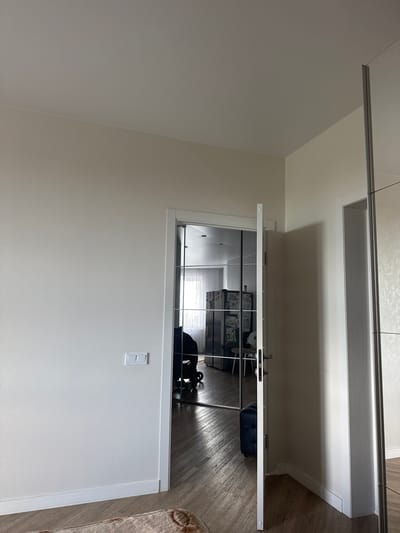The image shows a hallway with a closed sliding glass door. Through the glass, a room with a large black cabinet or wardrobe is visible. The cabinet has framed pictures or artwork on its doors. The room appears to be a bedroom or dressing room, with a black armchair and a dark bag on the floor. There are also some items on top of the cabinet that are difficult to discern.
The hallway itself has a light-colored textured wall with a white light switch. Above the sliding door, there is a narrow white shelf or ledge. The floor in the hallway is covered with wooden laminate flooring. The overall impression is of a modern, clean, and well-lit interior space. The time of day cannot be determined, and there are no visible weather cues. The location is Vsevolozhsk, Russia. No people are visible in the image. There is no visible text.
