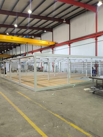 This image captures an interior view of a large industrial facility, likely a workshop or manufacturing plant, located within the Municipal Unit of Inofyta, Greece. The scene is dominated by a partially constructed modular structure, characterized by a white metal frame and a wooden floor. Several other modular units, some appearing complete and others in various stages of assembly, are visible in the background, along with ladders, scaffolding, and other construction materials.

Two individuals are present in the frame. One, wearing a light-colored shirt and dark pants, stands near the left side of the modular structure, seemingly engaged in work. Another individual, dressed in blue work attire, is positioned further in the background on the right, also appearing to be working.

Overhead, a prominent yellow crane beam with "10t" and "metalkat.eu" markings is suspended. The ceiling of the facility features exposed metal beams and industrial lighting. The floor is a grey concrete surface with distinct yellow painted lines, indicating pathways or work zones. Debris, including small pieces of white material and scattered debris, is visible on the floor, suggesting ongoing construction or a busy work environment. The overall impression is one of an active industrial setting focused on the production or assembly of modular buildings.