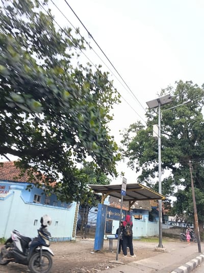 A blue and white building, possibly a school or public facility, stands behind a bus stop with two people. A motorbike is parked nearby. A solar-powered street lamp is beside the bus stop. Green trees and overhead wires frame the scene. A child with an adult are visible in the distance on the sidewalk.