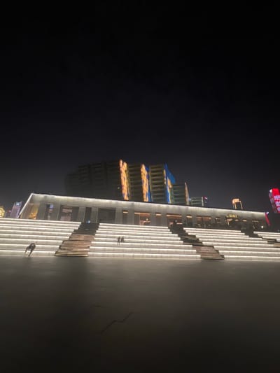 A wide-angle, low-light outdoor shot depicts the illuminated steps of a modern architectural structure at night. The steps are a prominent feature, uniformly lit with white LED strip lights embedded along the risers. These steps ascend towards a building with a contemporary facade, also illuminated with white and accent lighting. Behind this building, several taller structures rise into the dark sky, their upper portions displaying colorful, abstract light projections in shades of yellow, orange, and blue. A few individuals are visible on the steps; two people are in the lower left, appearing to walk together, while two more are seated further up on the right. The ground in front of the steps is a dark, paved surface, reflecting some of the ambient light. The sky is a deep black, suggesting nighttime. The scene appears to be in a public urban space, likely a plaza or outdoor venue. The textual signage on the building to the far right appears to be in Chinese, and the architectural style and lighting are consistent with modern Chinese cityscapes.