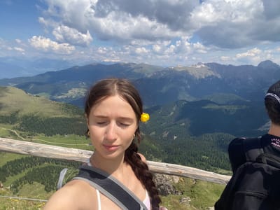 In a selfie taken from a mountain viewpoint in the Italian Alps, a young woman with closed eyes and a yellow flower tucked behind her ear is smiling. She is wearing a white tank top and a backpack. The background shows a vast expanse of mountain ranges, dense green forests, and a bright blue sky with white clouds. A wooden railing runs across the bottom of the image, with a glimpse of another person's head and backpack on the right side.
