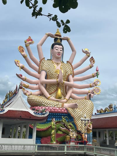 An outdoor, eye-level, full shot captures a large, multi-armed statue in Baan Plai Laem, Thailand, against a cloudy sky with green foliage framing the top. The statue, adorned in white and gold, sits cross-legged with hands in a prayer pose. Multiple arms extend from its torso, each holding a symbolic object, including a lotus flower, a wheel, a red orb, and a flame. Below the statue, a vibrant green dragon sculpture winds around its base. Two small human figures, one in green and one in yellow and blue, stand beside the dragon. In the background, traditional Thai temple architecture with red roofs and decorative elements is visible. The scene is serene, with no discernable action or interaction.