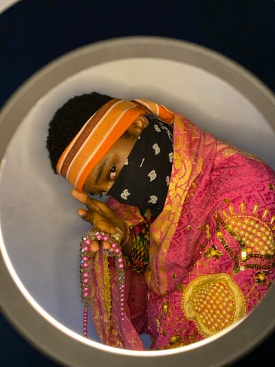 A single individual is positioned within the circular frame of a ring light, illuminating the scene against a light grey background with a darker upper edge. The person has short, dark, curly hair and wears a striped headband in orange, brown, and off-white. A black bandana with white patterns covers the lower face, revealing only dark eyes that gaze towards the viewer. The individual is draped in a vibrant hot pink garment, richly embroidered with gold metallic patterns and embellished with large, circular designs of white pearls or beads and scattered gold sequins on the visible left side of the chest/shoulder. Layers of other fabrics, including a multi-colored geometric pattern and gold-threaded material, are visible on the right arm area. The person's right hand is raised, holding multiple strands of beads, including prominent pink and clear faceted beads, as well as smaller gold beads. Bracelets are visible on the right wrist. The scene takes place in Ngleshi Amanfro, Ghana.
