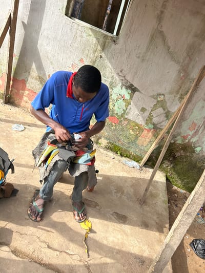 An outdoor scene in Ihiagwa, Nigeria shows a man sitting on a low stool and working on what appears to be shoes. He is wearing a blue t-shirt with a red collar and grey pants. The man is holding a white object, possibly glue or sealant, in his right hand and is manipulating dark-colored shoe material with his left hand and tools. The stool he is sitting on is covered with a patterned cloth, and he is wearing flip-flops. In front of him, on the ground, is a discarded banana peel. The background consists of a rough, weathered concrete wall with peeling paint in green, red, and white, and some moss growth. Two wooden poles lean against the wall. The ground is dirt and concrete.