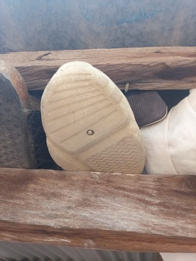 The image displays a close-up, overhead view of the sole of a worn, light-colored shoe resting on wooden planks. The sole exhibits a pattern of parallel lines and wavy ridges, with a small circular indentation at its center. To the left of the shoe sole is a portion of a dark, textured object, possibly another shoe or tire. To the right of the sole is a crumpled white fabric object and a dark gray, textured material. The background consists of wooden planks and a blurred, grayish surface. The scene appears to be in a dusty or outdoor environment, possibly a storage area or a discarded collection of items. The image does not provide any specific geographical markers to indicate its location in Zinna, Nigeria.