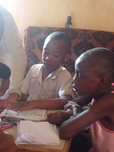 Two young children, possibly siblings, are sitting at a table covered with papers, books, and coloring supplies. One child wears a white collared shirt. The other child wears a pink tank top. An adult sits behind the child in the collared shirt. The environment is a simple interior, with a patterned couch and a bottle on top of the couch. The scene is likely taking place in a home in Yelwa, Nigeria.