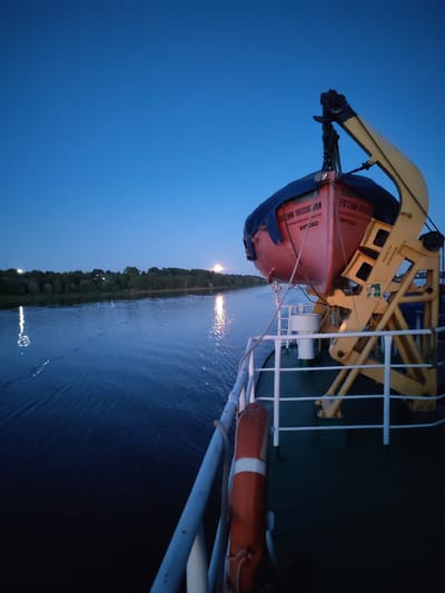 The image depicts the deck of a ship at twilight, with a clear blue sky and a calm body of water. On the deck, a red lifeboat is suspended by a yellow crane. The lifeboat has text in Russian, which appears to say "ЕВГЕНИЙ ОРЛОВСКИЙ" and "КОТЛАС". A life preserver is visible on the railing. In the distance, a tree-lined shore is illuminated by the fading light. The Volga River is a prominent feature of the landscape, reflecting the sky and distant lights. The ship appears to be docked or moving slowly along the river. The presence of the ship and the Volga River suggest a location in Yaroslavl, Russia, a city situated on the Volga.