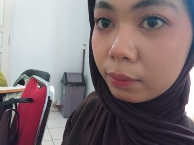 A close-up selfie shows a young woman wearing a dark brown hijab. She is facing the camera, and the composition focuses on her face, particularly her eye and lips. The background is blurred, but hints of an indoor environment are visible. To the left, a portion of a red chair with a chrome frame is in the foreground. Further back, a grey trash can is visible against a white wall. The scene appears to be indoors, possibly in an office or public space.