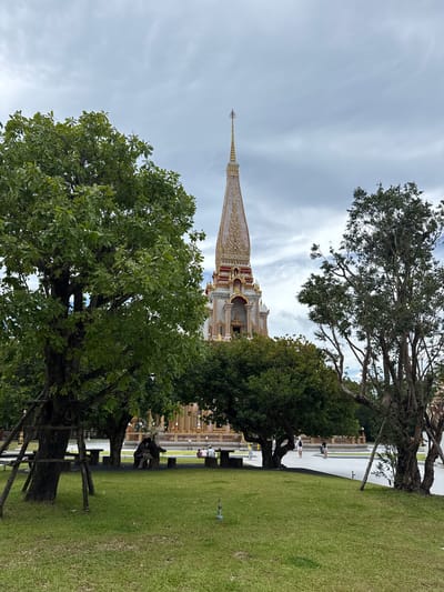In Chalong, Thailand, a serene temple complex is captured under a cloudy sky. In the foreground, a lush green lawn is dotted with park benches and picnic tables. Two individuals are seated at one of the tables, facing away from the camera, appearing to engage in conversation. To the left and right of the frame, mature trees with dense foliage provide shade. Behind the trees, the ornate spires of a Buddhist temple rise majestically. The temple's facade features intricate golden detailing and red accents against a pale structure. In the mid-ground, several other people are visible walking around the temple grounds. The scene conveys a sense of tranquility and cultural significance.