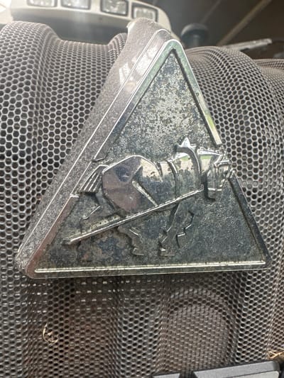 A triangular chrome emblem featuring a horse pulling a plow is mounted on a textured, honeycomb-patterned surface. The emblem shows signs of wear and dirt, with the chrome finish scratched and tarnished in places. The honeycomb surface is also dusty. The background is out of focus but appears to be a part of a vehicle, possibly a tractor, with hints of lights visible at the top. The scene is lit by natural light. The image was taken outdoors, likely in an open area given the context of agricultural machinery. There are no people or animals present in the image.
