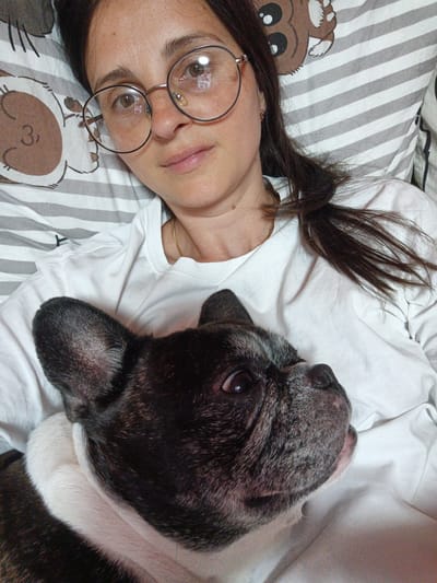 A woman wearing round glasses and a white t-shirt lies in bed with a French Bulldog. The woman has dark hair and is looking towards the camera with a neutral expression. The dog is black and white and is nestled against her chest, looking to the side with its mouth slightly open. The bedding has a pattern of gray and white stripes, with a graphic of a bear face on the pillow. The scene appears to be indoors, likely in a bedroom. The lighting suggests it could be daytime. There is no visible text in the image.