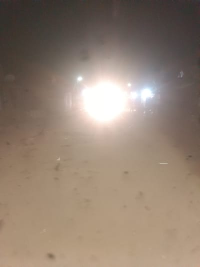 The image shows a dimly lit street scene at night in Wukari, Nigeria. A bright light from an unknown source, possibly headlights or a strong spotlight, dominates the center of the frame, creating a glare and obscuring details of the surroundings. The street appears to be unpaved, with a dusty or sandy surface. Several indistinct shapes suggest the presence of vehicles or structures in the background, further blurred by the bright light and possibly a low-resolution camera or poor lighting conditions. There are no clearly identifiable people or specific objects, only general forms and the overwhelming illumination.