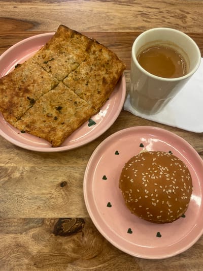 On a wooden surface, possibly a table, is a mug of tea, a plate of paratha, and a plate with a sesame seed bun. The paratha is cut into sections. All three are on pink plates decorated with small green hearts. A white napkin is near the mug. Given the food items, this may be a cafe in Mumbai, India.