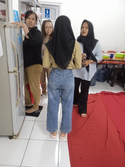 The image shows four women indoors. One stands next to a refrigerator, dressed in a black top and tan pants, and is barefoot. Another woman, wearing glasses and a striped shirt, stands behind her. The third woman, whose back is to the camera, is wearing a tan shirt, blue jeans, and a black hijab. The fourth woman is wearing a black hijab, a light blue top, and black pants. The floor is tiled and partially covered by a red sheet. In the background, there is a table with water bottles and containers, and a door with a restroom sign is visible. The location is inferred to be in Semarang, Indonesia, based on cultural cues and context of the image, such as the hijab worn by some of the women. The women are interacting in an informal setting, possibly a breakroom.