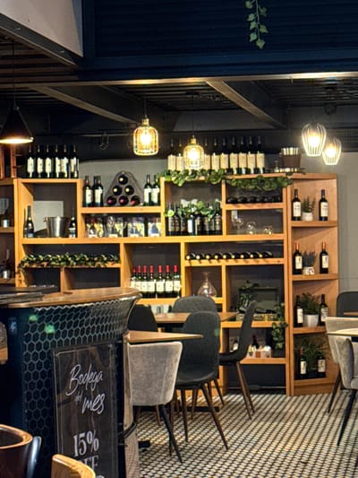 A restaurant interior showcases shelves packed with wine bottles, accented by decorative greenery. The shelves are part of a wooden structure, possibly a wine display. In front of the shelves, several chairs with wooden legs are stacked, one featuring a visible 'Bodega del Mes 15%' sign on its back. The room is illuminated by industrial-style pendant lights.