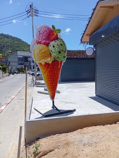 An oversized, flat cutout of an ice cream cone with four scoops of ice cream—strawberry, vanilla, mint chocolate chip, and another yellow flavor—is positioned on a sidewalk in Baan Lamai, Thailand. The ice cream cone is mounted on a black metal stand and rests on a gray concrete base. Behind the ice cream cone, a building with a thatched roof and gray metal roll-up doors is visible. The street in front of the building is paved, with a sidewalk made of patterned gray tiles. A few cars and motorcycles are on the street, suggesting it is a public area. In the background, a lush green hill rises under a clear blue sky with scattered clouds, indicating a tropical or sub-tropical environment. Electrical wires and poles are present along the street.