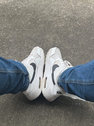 The image shows a view of a pair of white Nike Air sneakers and denim jeans on an asphalt surface. The shoes are slightly dirty. The location is indicated as Edison, United States of America, but there are no specific details in the image to confirm this.
