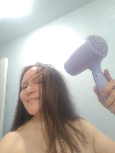 A woman is holding a light purple hair dryer in her right hand and drying her brown hair in a bathroom. She is smiling slightly with her eyes closed. The bathroom has light blue or gray walls and a white door. There is a bright light in the ceiling. The city of Neftekamsk, Russia is not discernible from the image.
