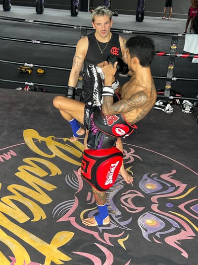 Two men are in a Muay Thai boxing ring. The man on the left has light-colored hair and a mustache, wearing a black tank top and black boxing shorts with a gold chain. The man on the right is shirtless, showing off a tattooed arm and torso, wearing black boxing shorts and holding kickboxing pads. He is actively training the man on the left. The gym floor has a large graphic of a stylized face and lettering. Visible text includes "TWINS SPECIAL," "MUAY THAI," and "BANGKOK." The scene appears to be indoors, likely during the day, given the lighting. The overall atmosphere suggests a training session in a martial arts gym. The location context is Choeng Thale, Thailand.